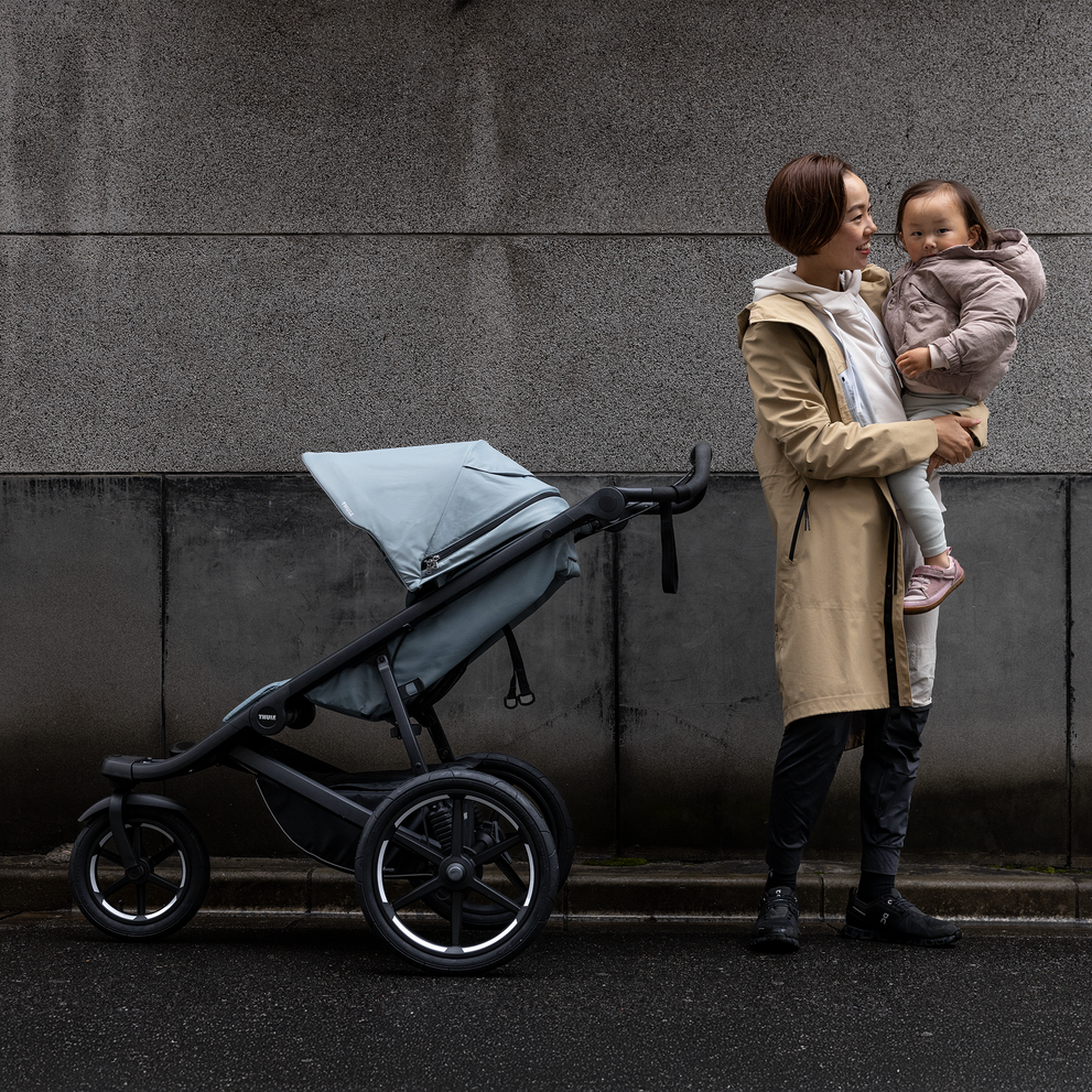 Parent holding child next to parked Thule Urban Glide 3 stroller on rainy street.