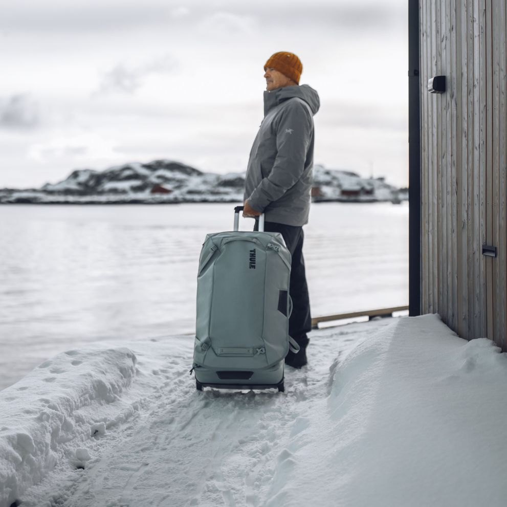 A man stands looking over icy ocean landscape with a blue Thule Chasm check-in suitcase.
