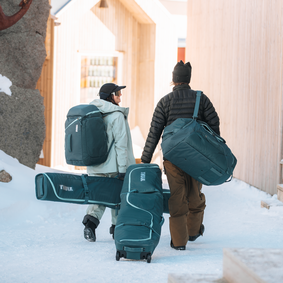 Two men are walking in the snow with their ski gear in a Thule RoundTrip Ski and Snowboard Boot Backpack and a Thule RoundTrip Ski and Snowboard Boot Backpack Duffel.