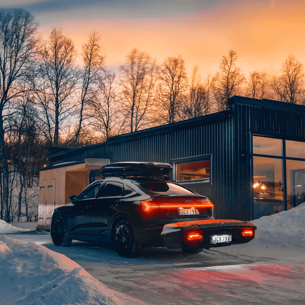 A sleek black SUV with Thule Arcos XL mounted, is parked outside a modern cabin at dusk, with snow on the ground and warm light glowing from the windows.