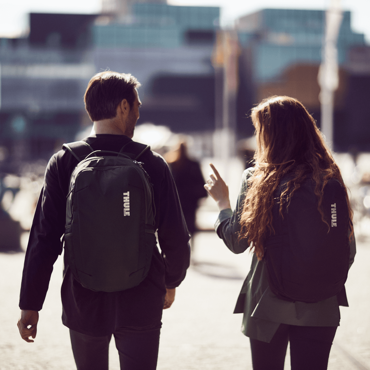 Two people walk on a city square with glass buildings in the background, holding Thule Subterra backpacks.
