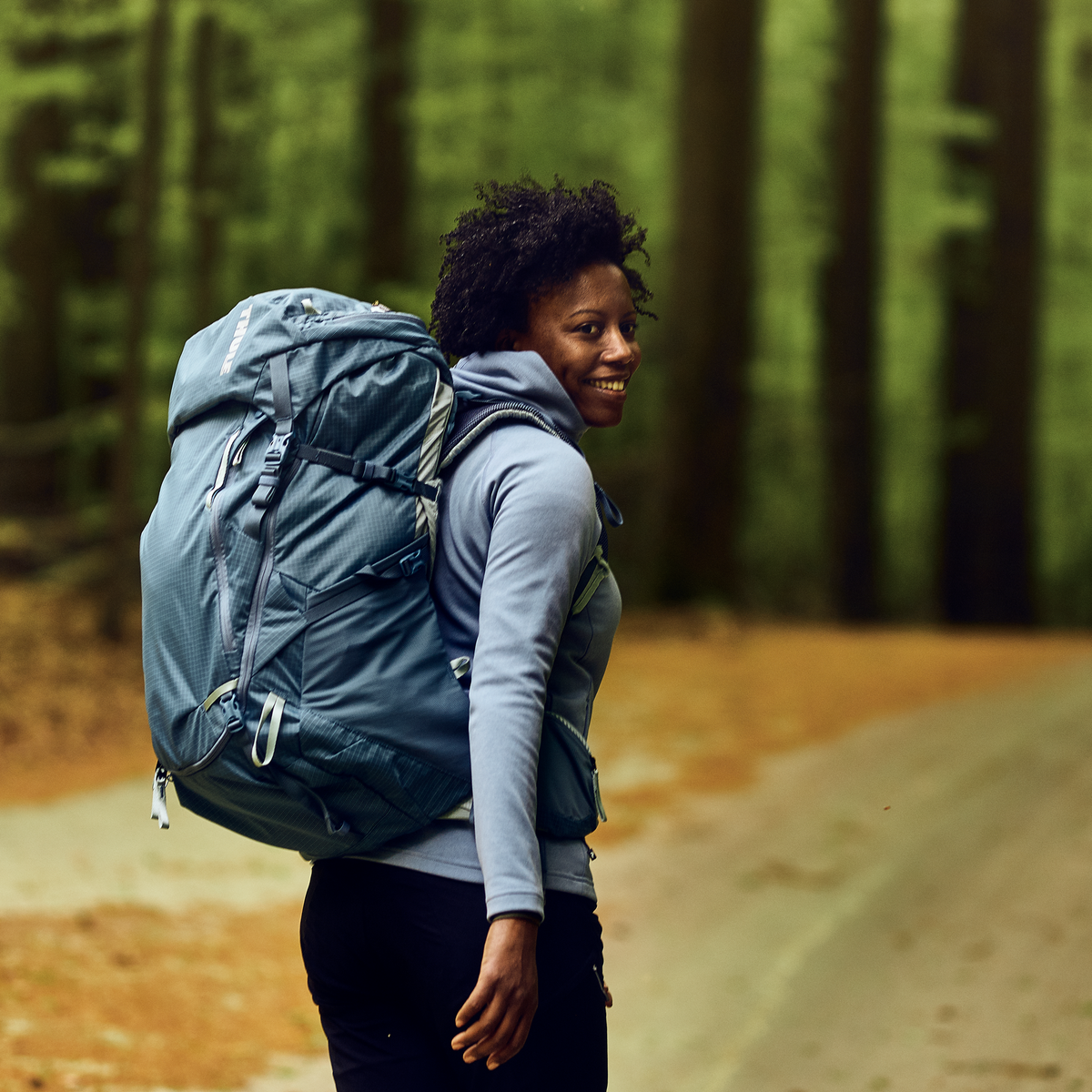 A woman walks down a forest road carrying a Thule Versant 60L hiking backpack.