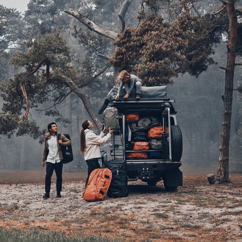 In a forest, a child stands on top of a jeep and unloads Thule Chasm duffel bags and hands them to his mother.