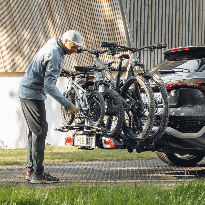 A man in casual outdoor clothing loads 4 bikes onto a bike rack attached to the back of an SUV in a sunny setting, in a covered parking area.