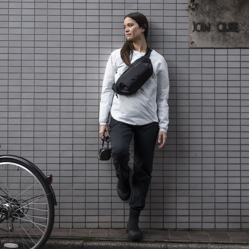 A woman leans against a brick wall carrying a black Thule Subterra crossbody sling.