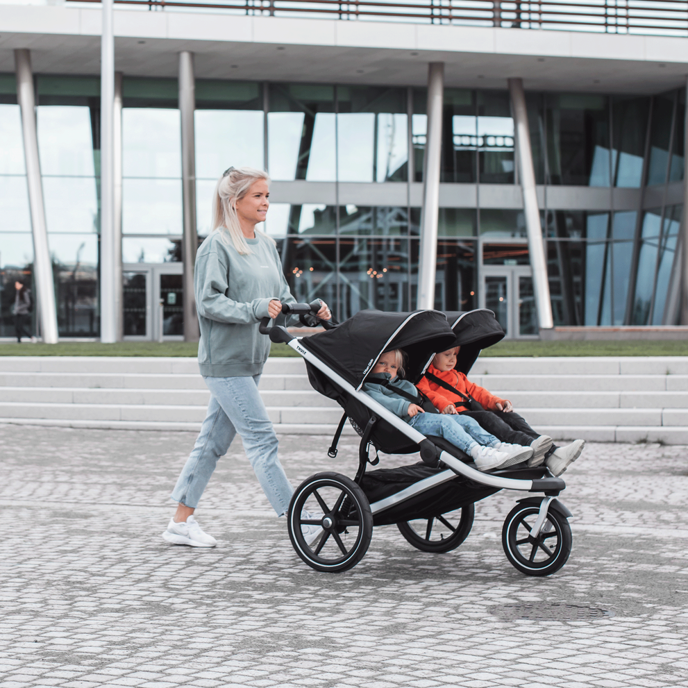 A woman walks down a cobbled street with her kids in a Thule Urban Glide 2 double jogging stroller.