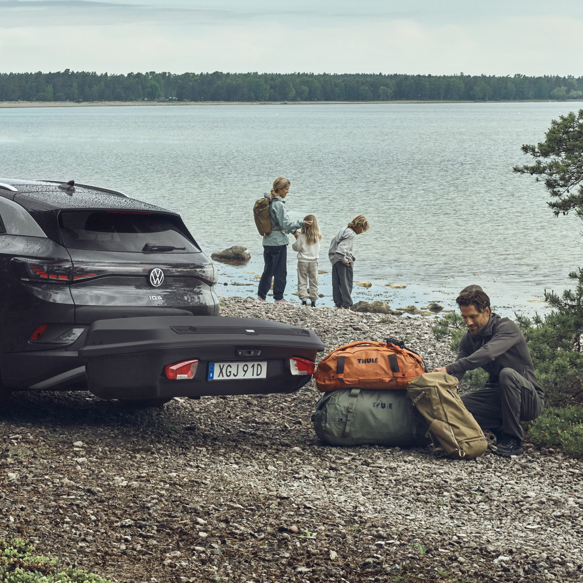 A family is standing beside their car by a lake. A man is unloading bags from a Thule Arcos cargo box.