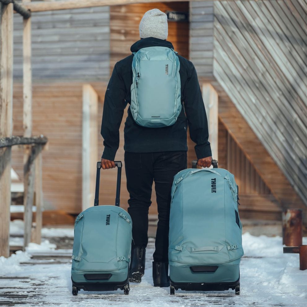 A man is walking down a snowy path with blue Thule Chasm suitcases and a backpack.