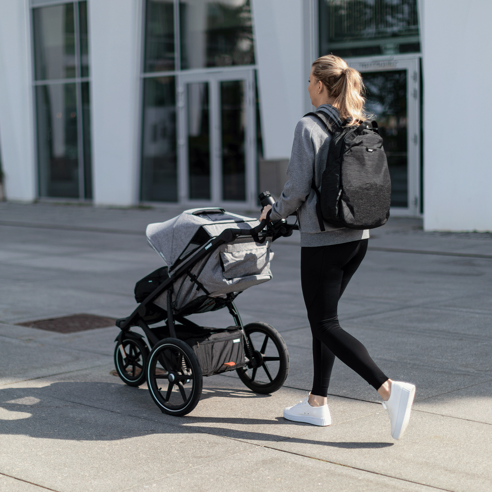 A woman walks down the street with her child in a gray Thule Urban Glide 2 jogging stroller.
