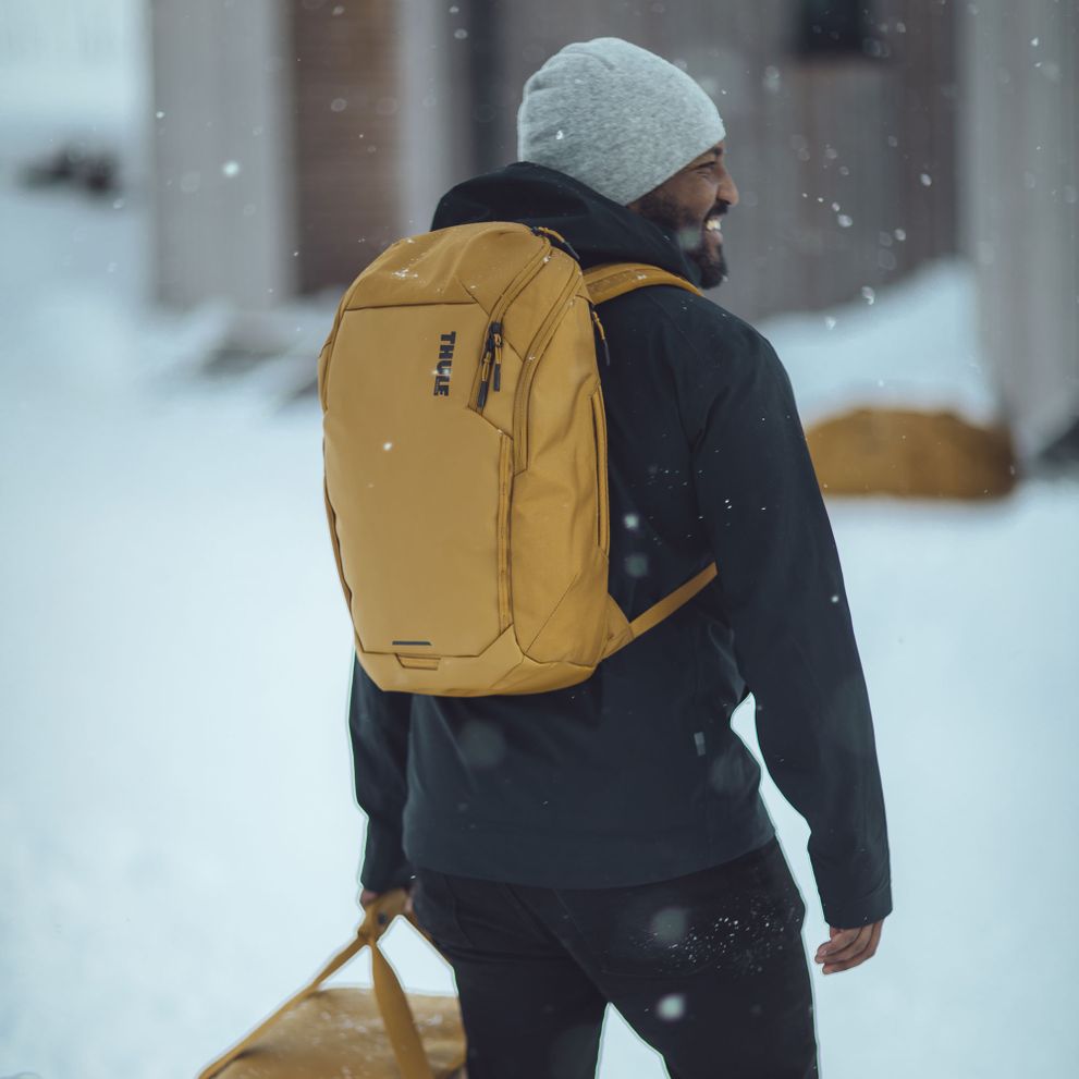 A man is standing in the snow with a beenie, and a orange Thule Chasm backpack.