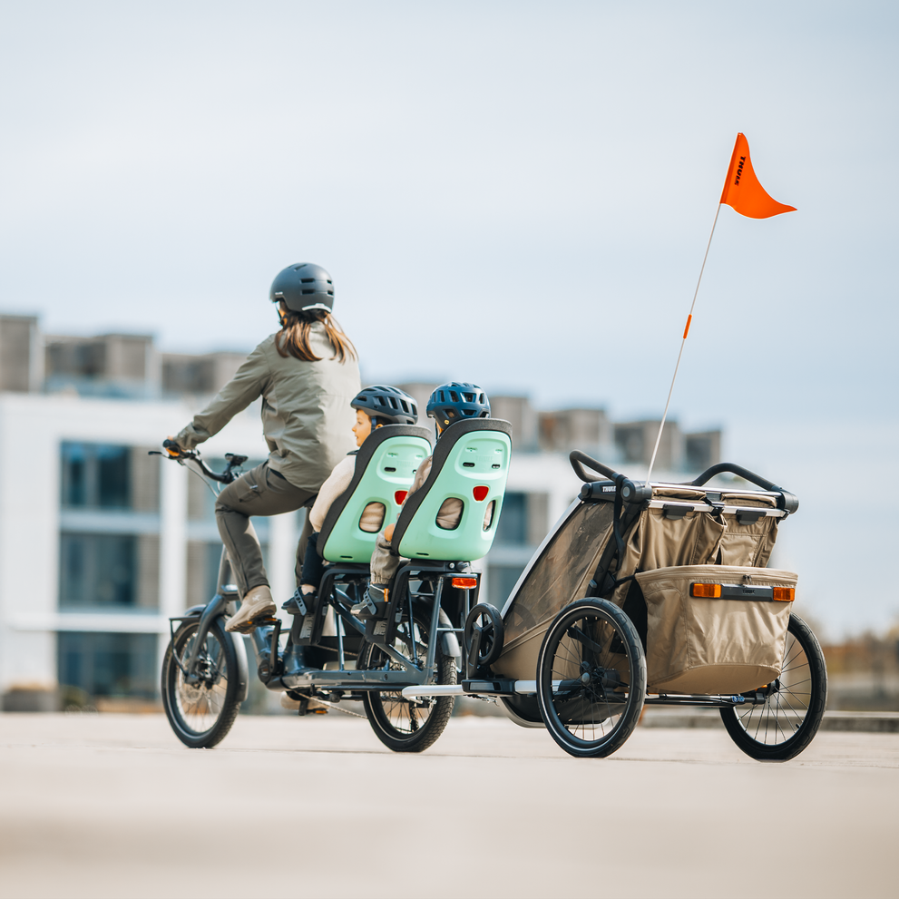 Two children sit in Thule Yepp Nexxt 2 seats on a bike, with a Thule trailer hitched behind.