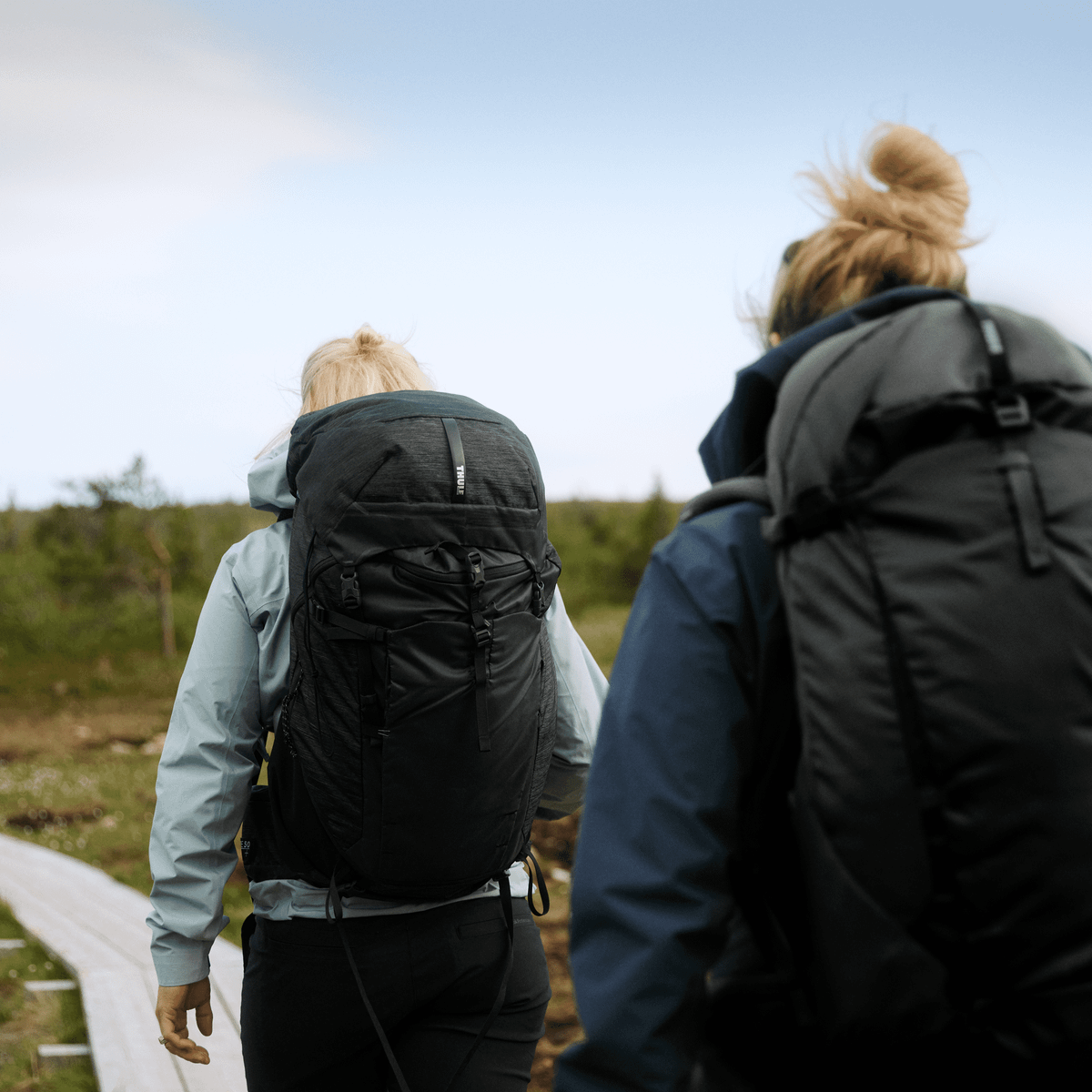 Two women walk down a country path carrying black Thule Topio 40L hiking backpacks.