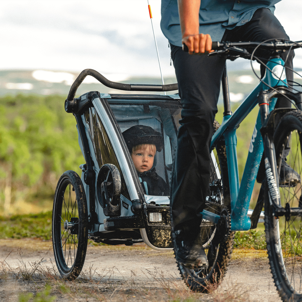 In a field, a man bikes with his blue Thule Chariot Cross child bike trailer.