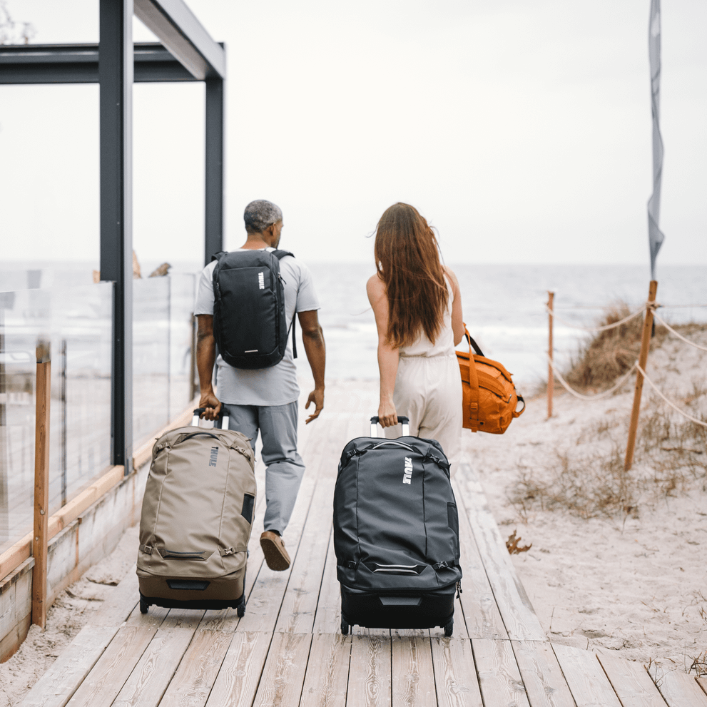 A man and woman walk down a wooden path next to a beach with black and green Thule Chasm check-in suitcases.