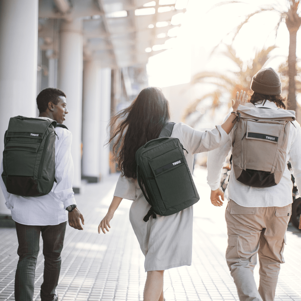 Three friends walk down a sunny street with palm trees, carrying Thule Paramount Backpacks.