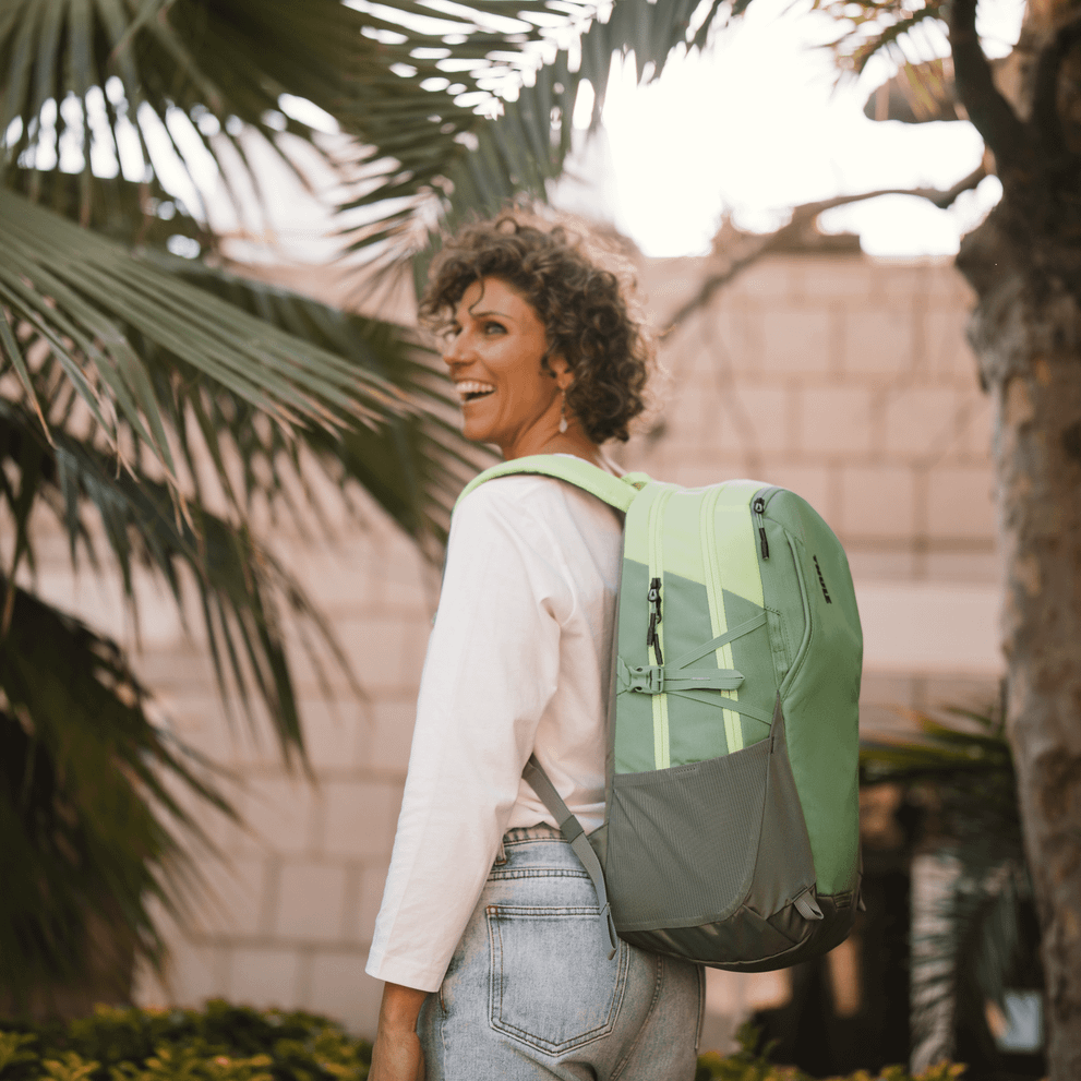 A woman stands next to a tiled building and a palm tree with a green Thule EnRoute backpack.