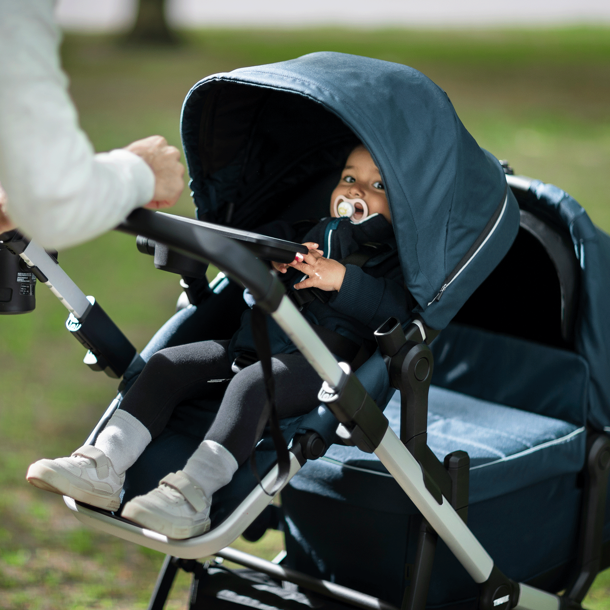 A close-up of a baby with a pacifier sitting inside the blue Thule Sleek double stroller.