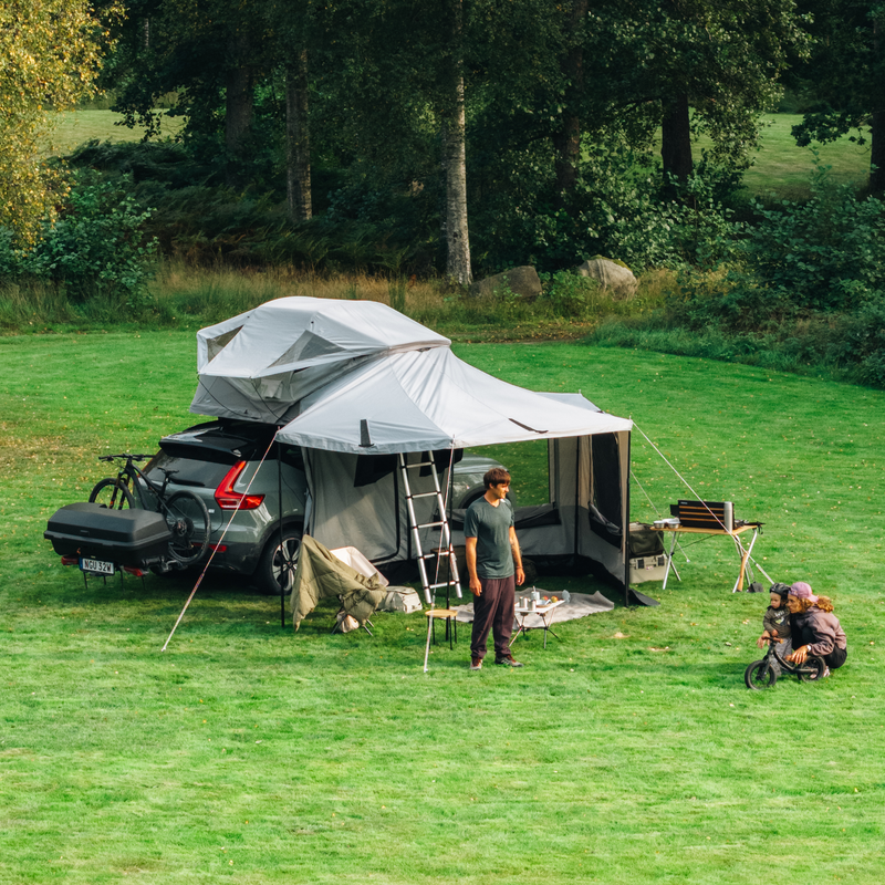 Thule Approach 2 soft-shell rooftop tent unfolded and mounted with awning on the top of a carr with one bike and Thule Santu cargo box on the towbar bike rack, parked in an outdoor setting and a family interacting by the car.