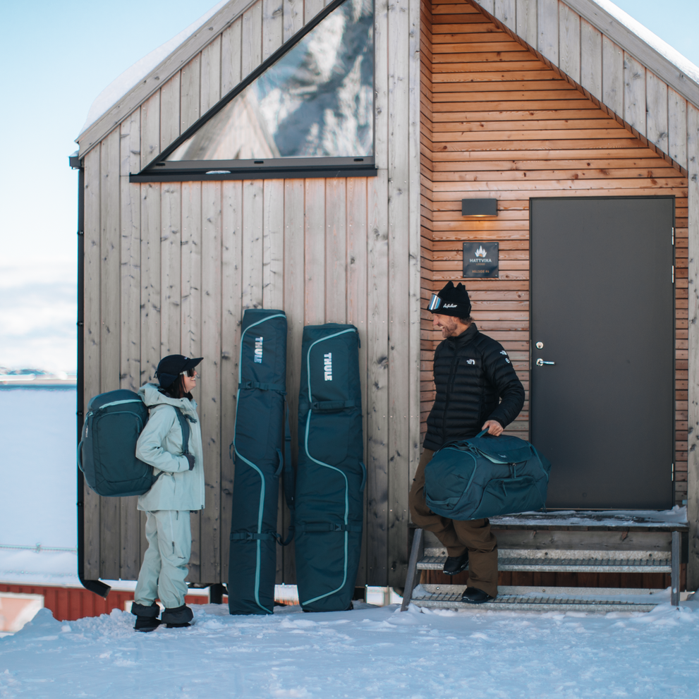 Two men are standing outside a wooden house with their ski gear in Thule RoundTrip bags.