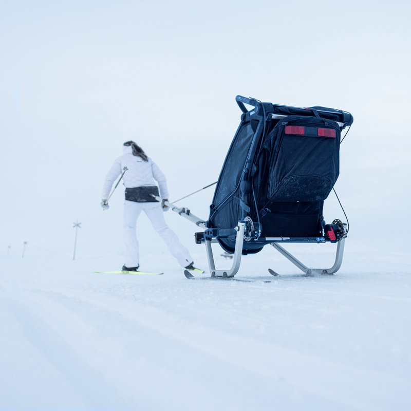 A woman cross-country skiing through a snowy landscape, pulling a Thule Chariot Sport 2 single, seen from behind.