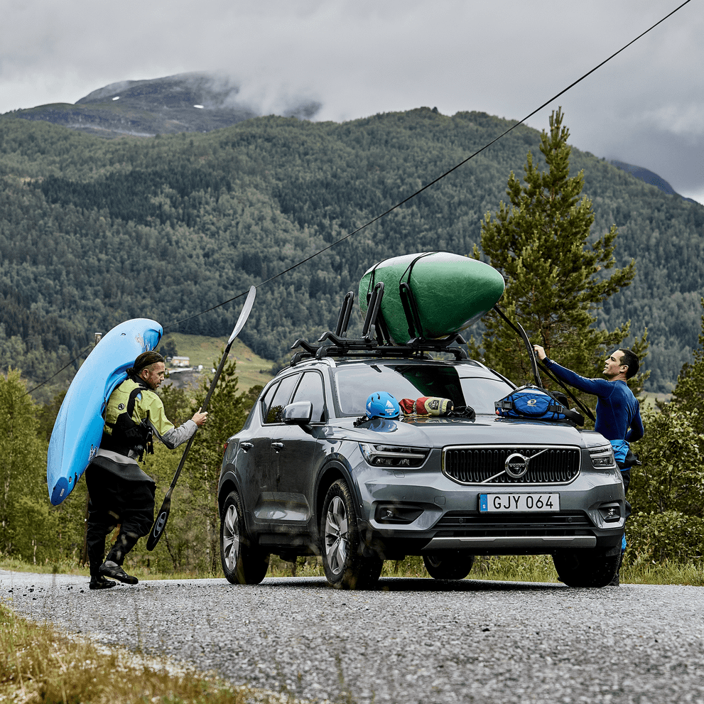 A man is carrying a kayak towards a car where anonther man is loading his kayak on the car roof with a Thule Hull-a-Port XTR