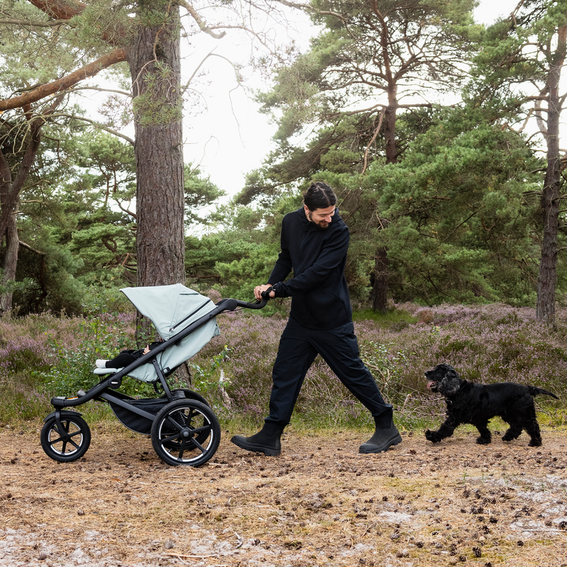 Mujer empujando una carriola Thule Urban Glide 3 por un sendero forestal con un perro.