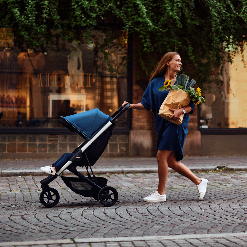 A woman in blue walks down a cobbled street with a blue Thule Spring compact stroller, holding groceries.
