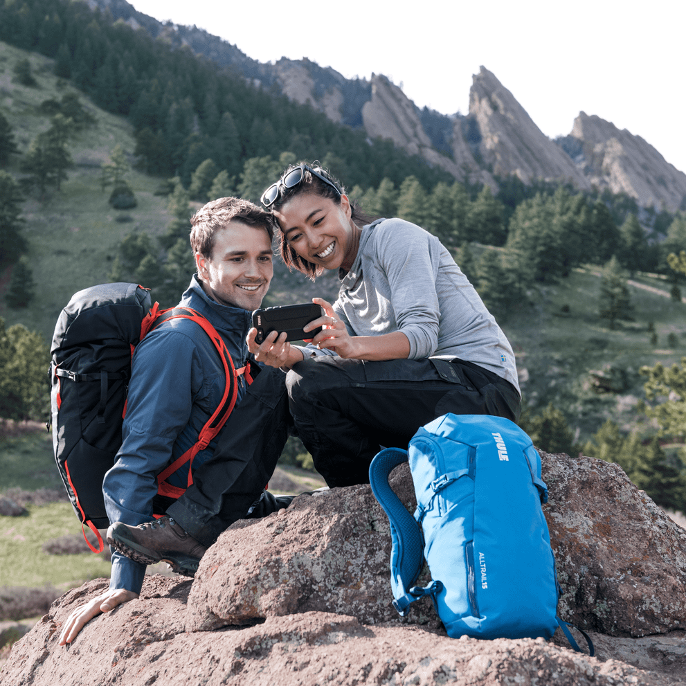 Two people sit in the mountains with Thule AllTrail 15L backpacks, looking at their phone.
