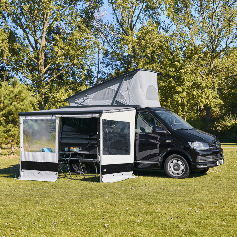 Compact van with pop-up roof and open Thule awning with panel set set up with a door opening on a grass field in the sun.