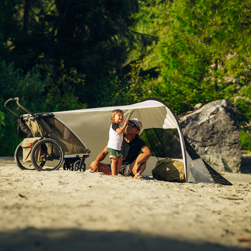 A man sitting and a child standing under the Thule sun and wind tarp, mounted on a Thule Chariot stroller.