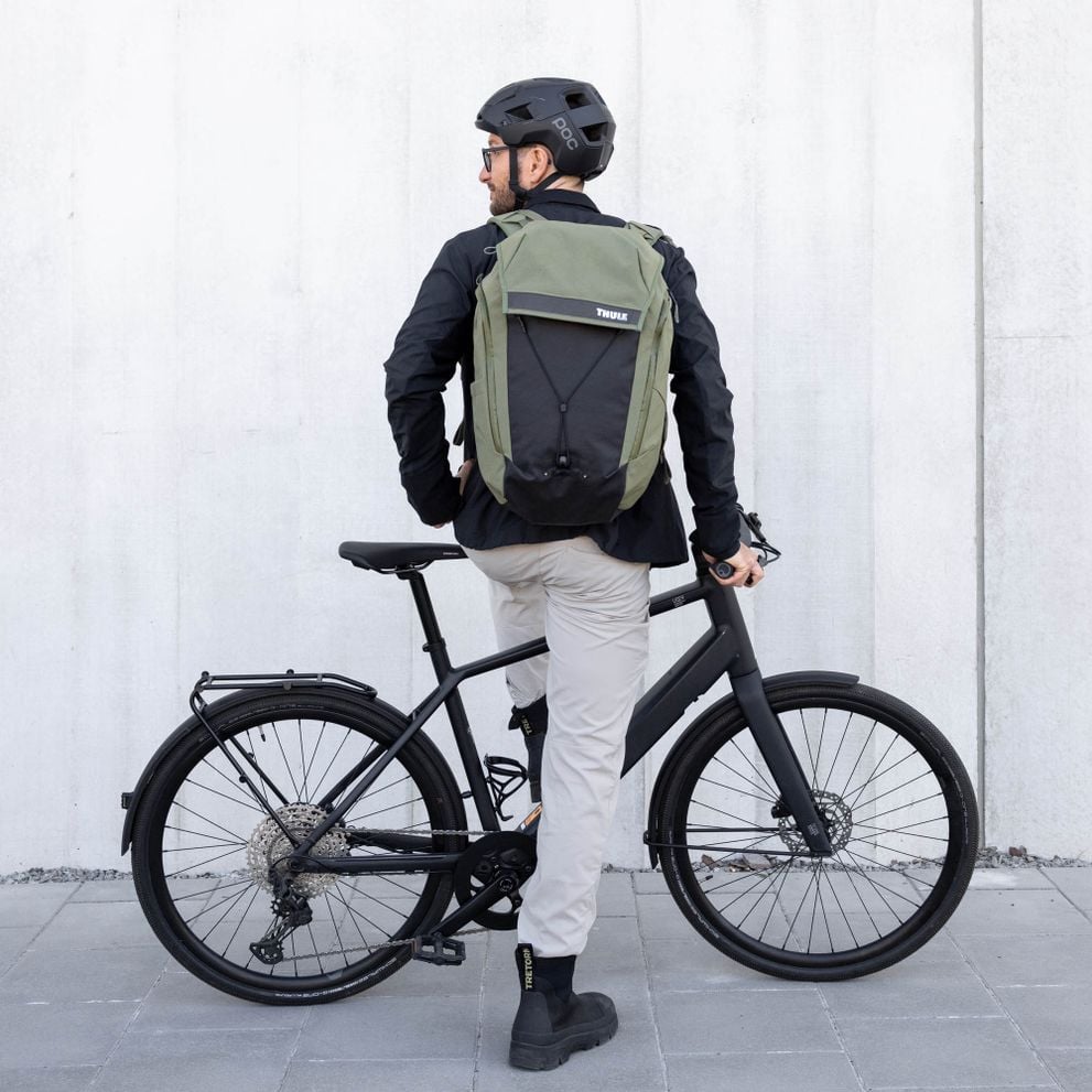 A man wearing a black helmet and jacket stands with his bike with his back towards the camera, carrying a green backpack, facing a concrete wall.