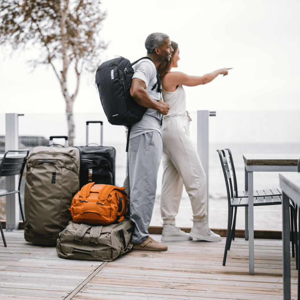 A couple stand and look out at the ocean with a pile of Thule Chasm duffels and suitcases beside them.