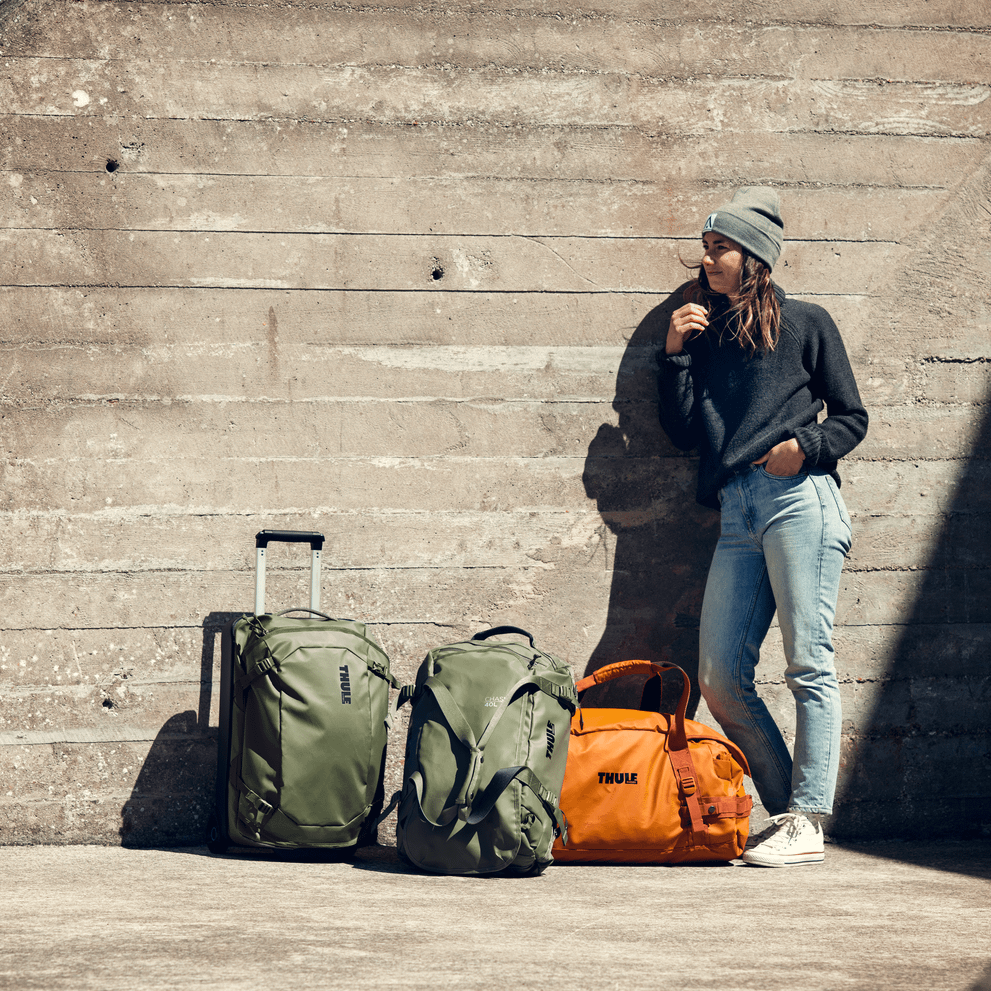 A woman leans against a concrete wall in the sun with a green Thule Chasm Carry On and two duffel bags.