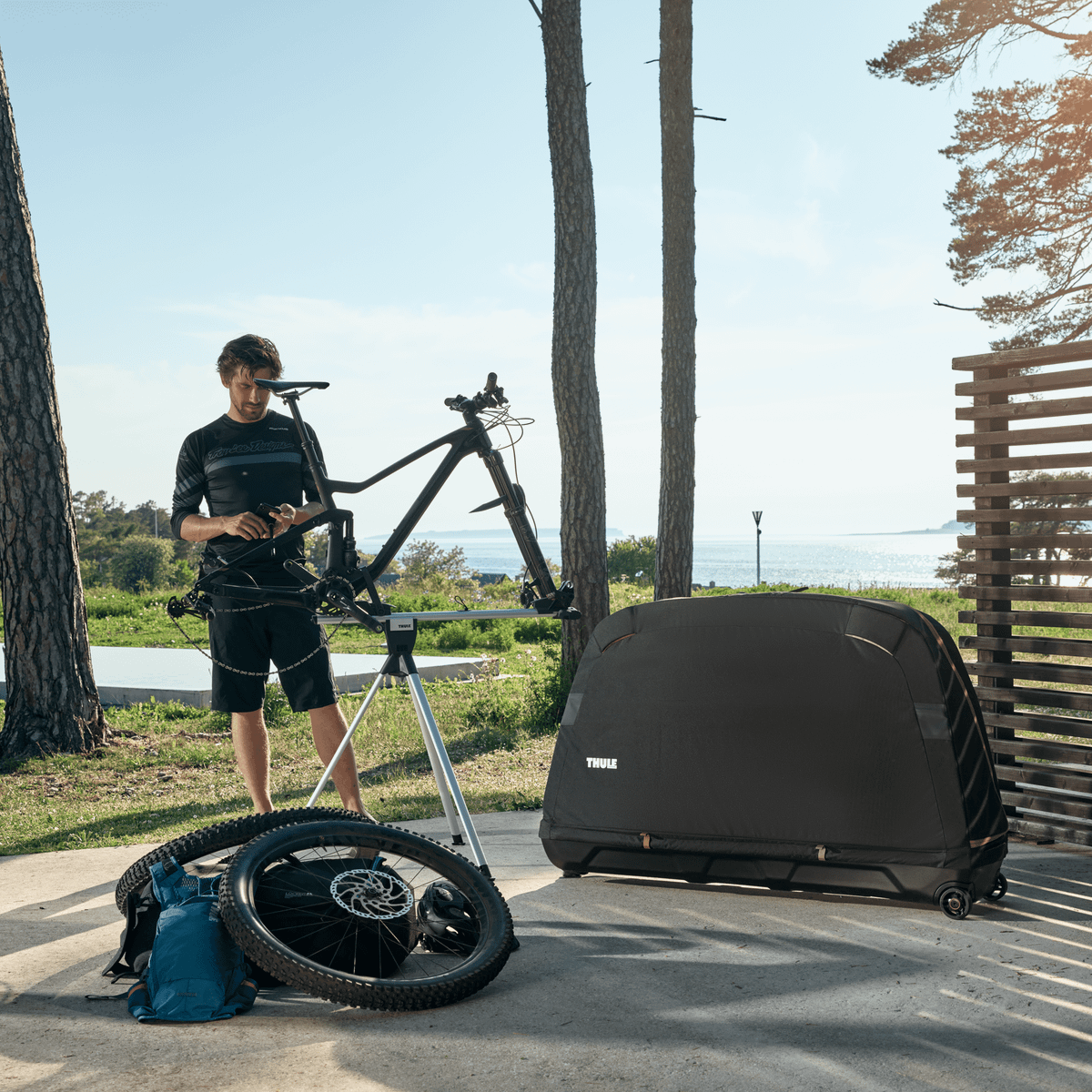 On a sunny outdoor area a man fixes his bike on a stand next to a Thule Roundtrip MTB bike travel case.