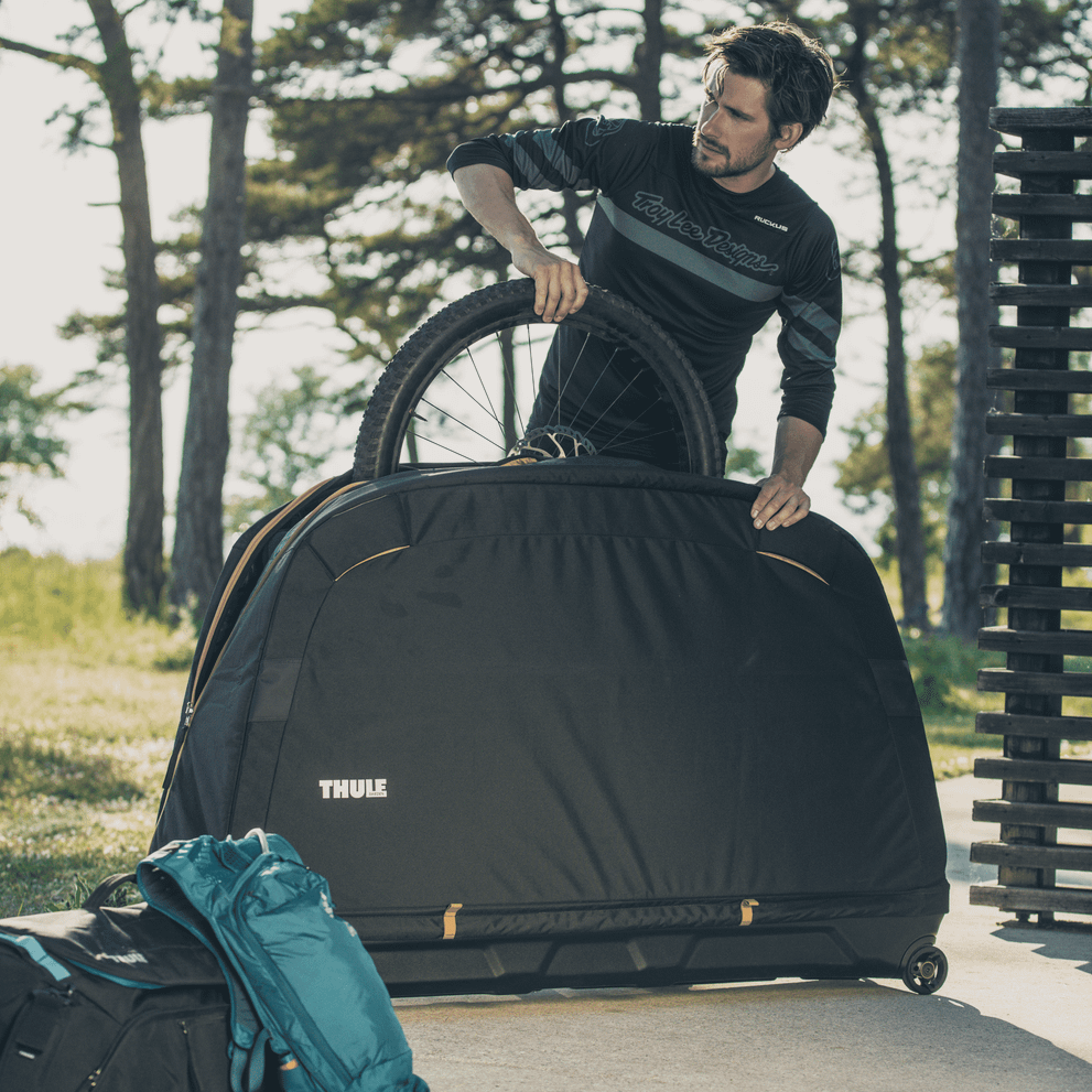 在树木之间，一名男子从黑色 Thule Roundtrip MTB 自行车旅行箱中取出一个轮子。