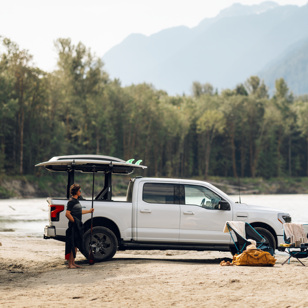 A person stands next to a pickup truck with a Thule Xscape truck rack with a roof box and a SUP board, at the beach.