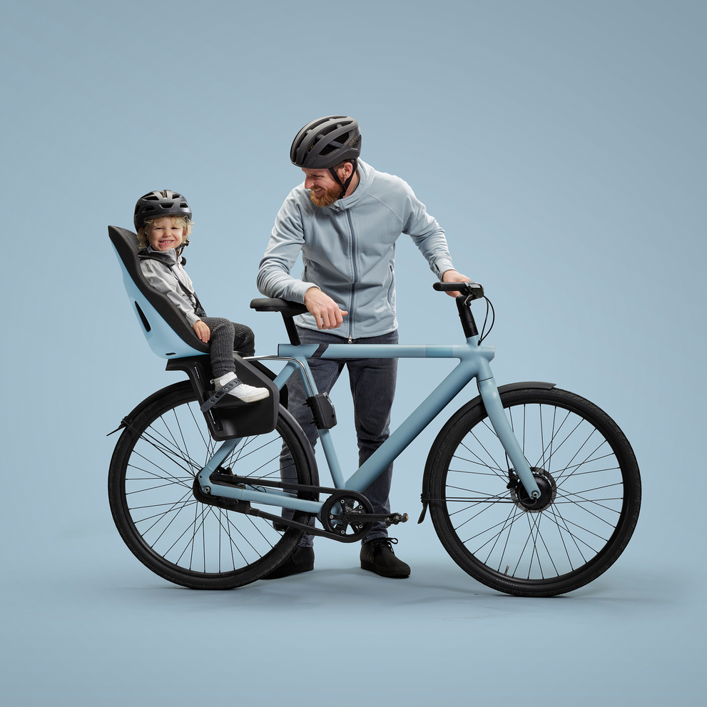 A smiling child, securely strapped into a light blue child's bike seat mounted on the back of a matching blue bicycle, looks on happily. A bearded man in a grey hoodie and black helmet stands by the bicycle
