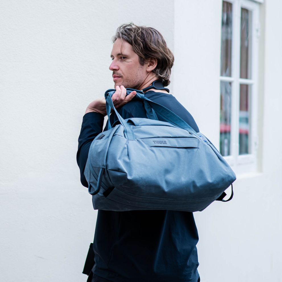 A man wearing a black coat holds a blue Thule Aion duffel over his shoulder.