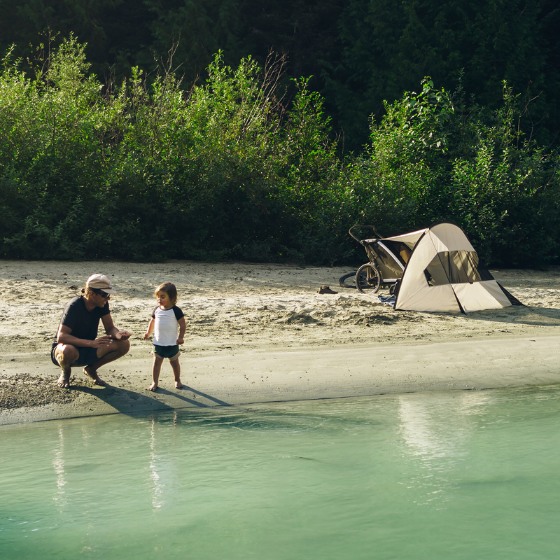 A man squatting and a child standing by the lake, with a Thule sun and wind tarp mounted on a Thule Chariot stroller in the background.