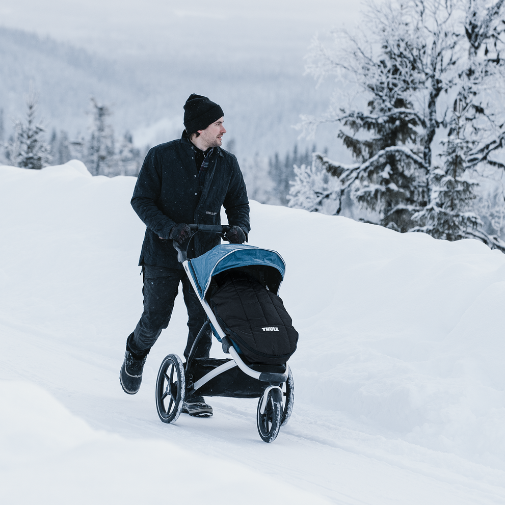 A man walks through the snow with his baby in a jogging stroller with a Thule Footmuff Sport.