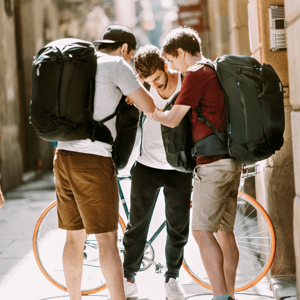 Three backpackers look at a map in a narrow sunny city street wearing Thule Landmark 40L travel backpacks.