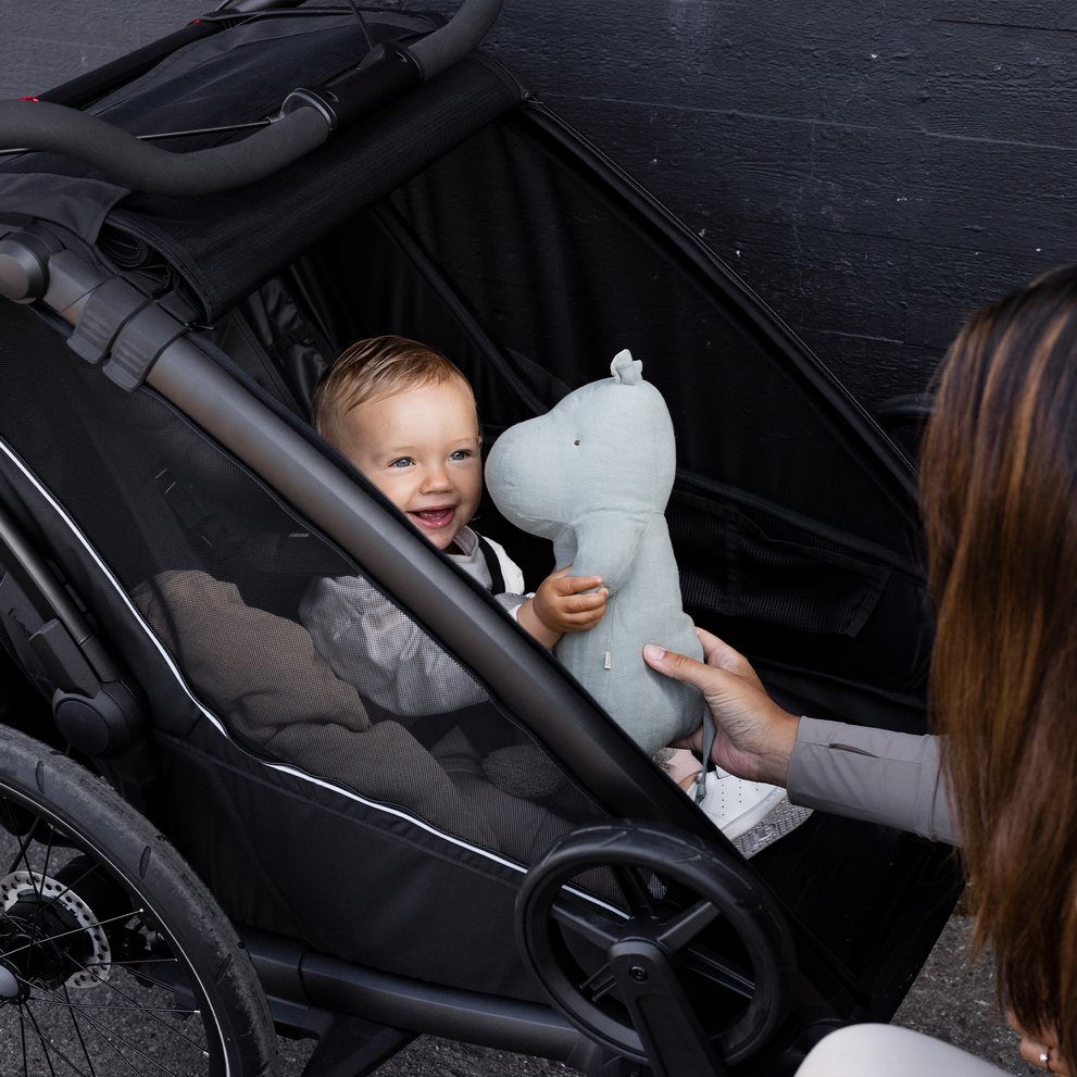 A baby sits in a black Thule Chariot Sport 2 bike trailer for kids holding a stuffed toy.