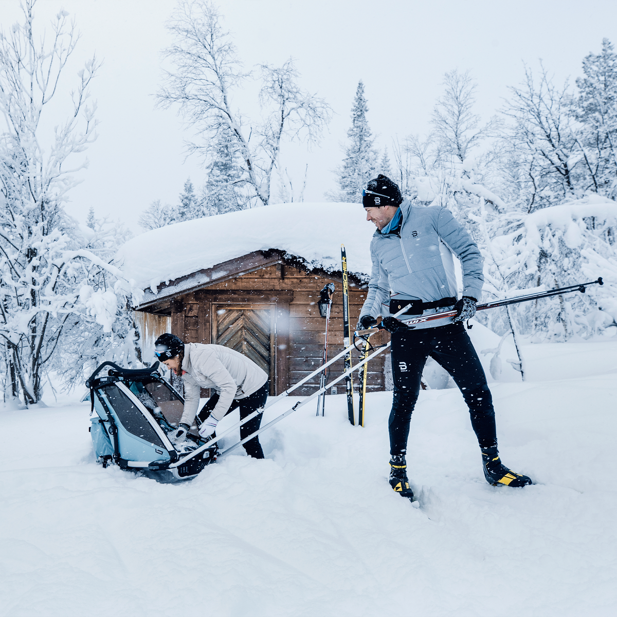 Outside a cottage, a couple goes cross country skiing with a kids' bike trailer using the Thule Chariot Ski Kit.