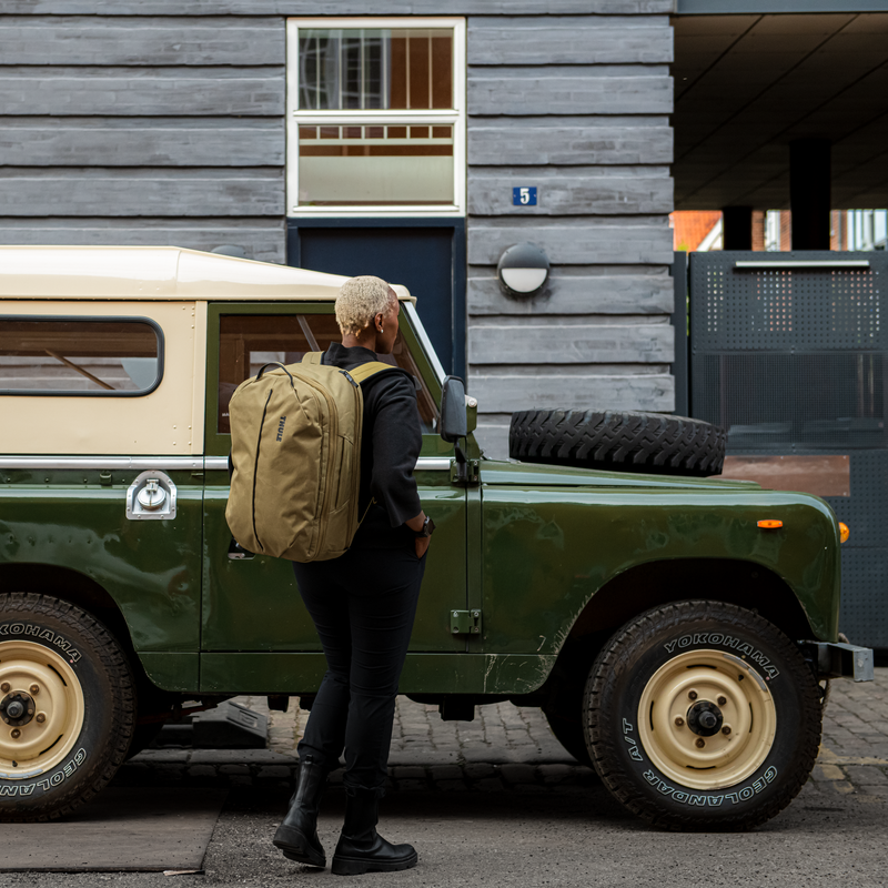 Uma mulher está ao lado de um carro verde carregando uma mochila de viagem Thule Aion bege.