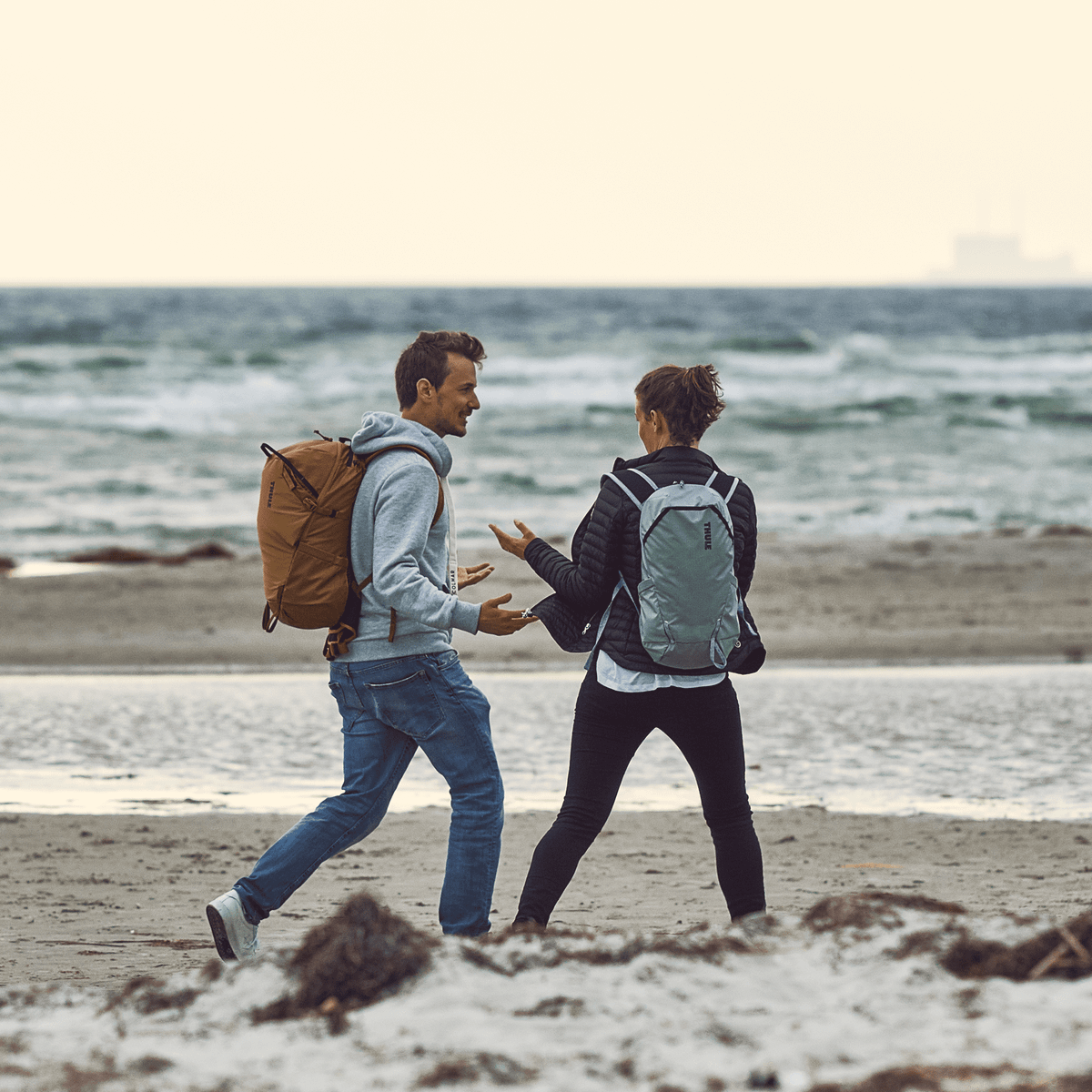 Two people stand and chat on a beach carrying yellow and blue Thule Stir 25L backpacks.