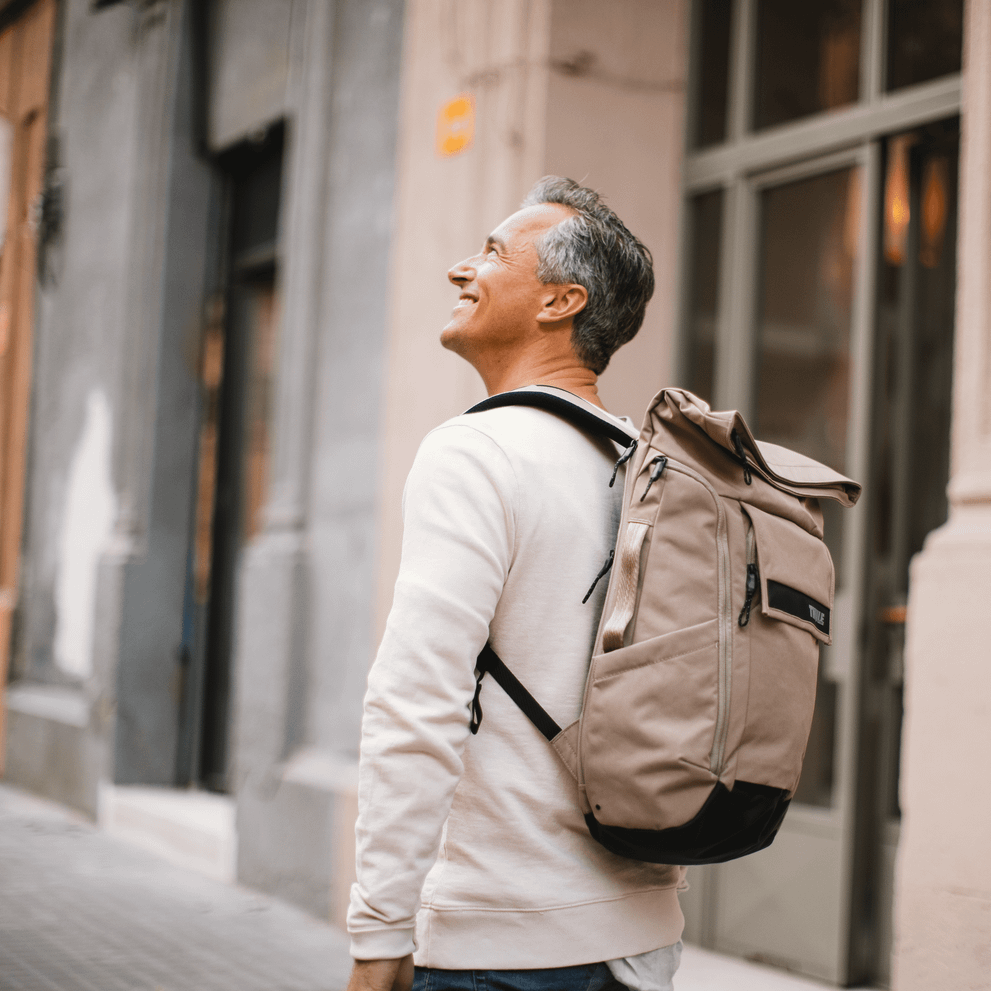 A man stands by a building and looks up, carrying a tan-colored Thule Paramount Backpack.