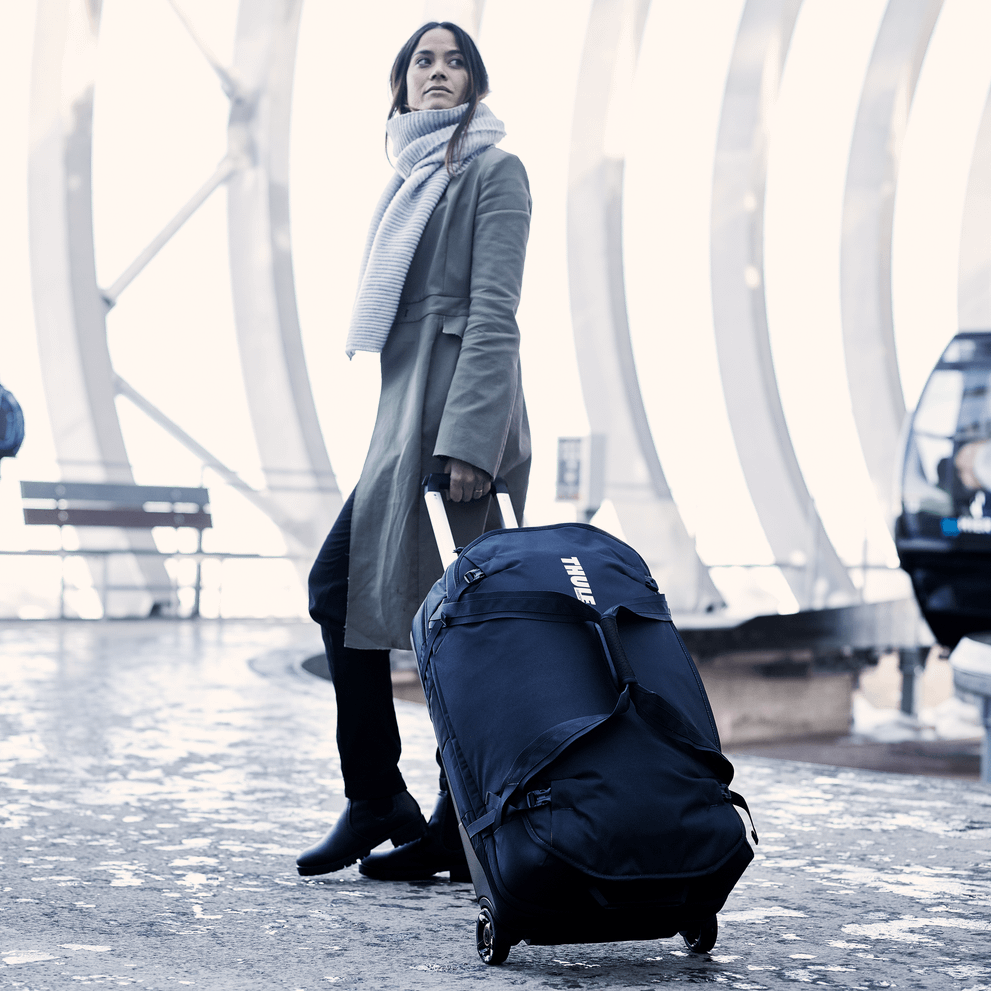 In a cold bus station, a woman waits with a scarf and her Thule Subterra Wheeled Duffel suitcases.