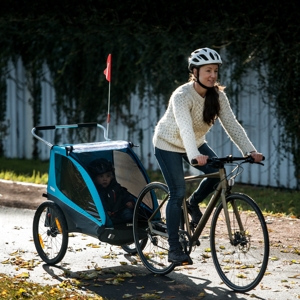 A woman bikes down a bike path with a blue Thule Coaster XT kids' bike trailer.