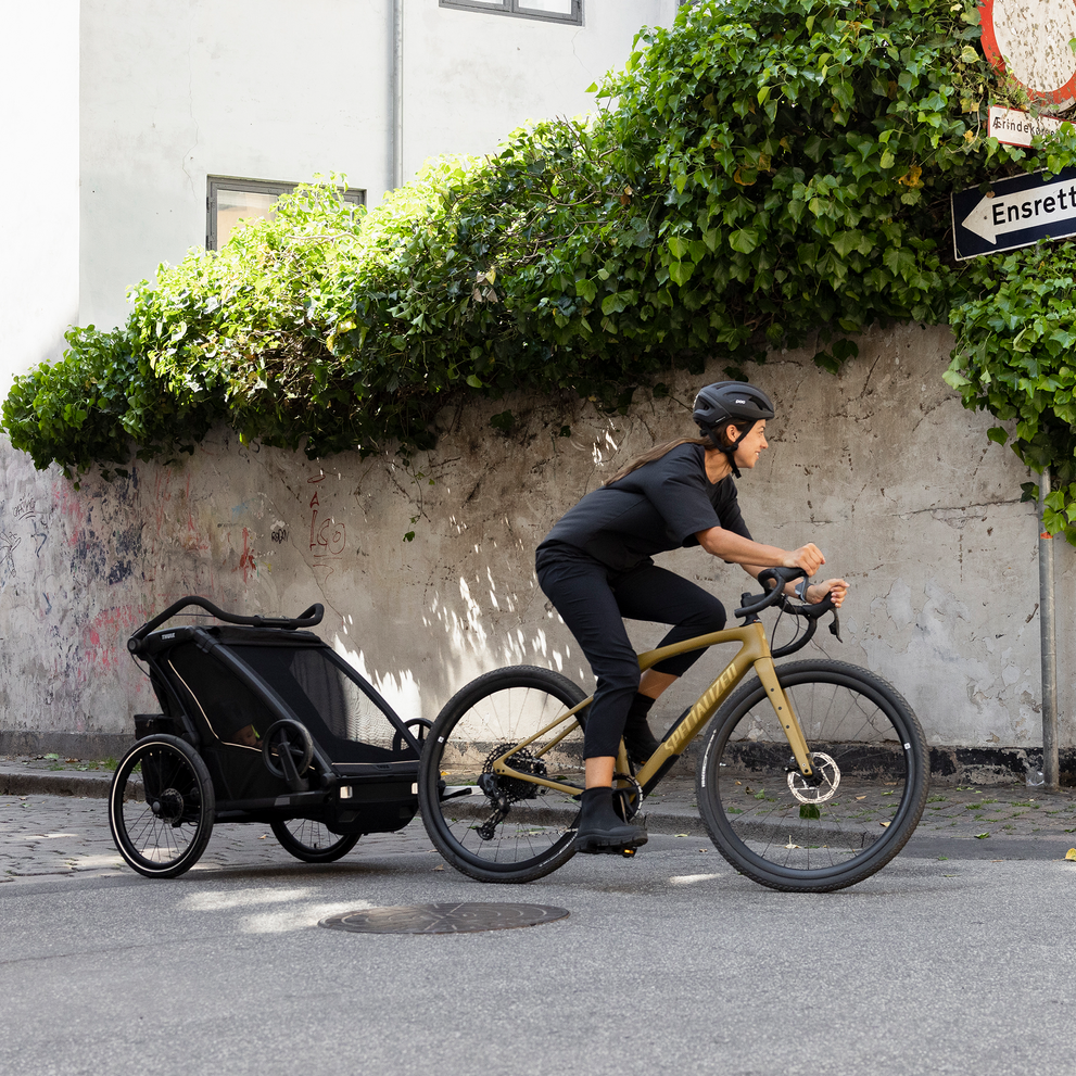 A woman cycles down a sunny street with her black Thule Chariot Sport 2 kids bike trailer.