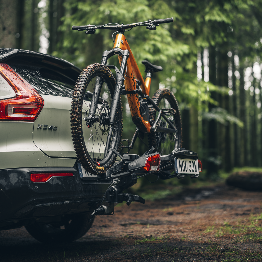 A closeup of a mountain bike on a Thule Outpace towbar bike carrier mounted on a sage green car, parked in the woods.